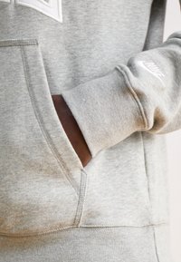 Sivi pulover s kengursko žepom, rebrastimi manšetami in belim Nike logotipom. Material izgleda mehak, s teksturirano flisasto površino.