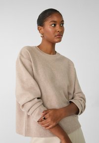 Beige gebreide trui met een ronde hals, geribde mouwen en een relaxte pasvorm. Glad textuur, met omgeslagen mouwen. Geen patronen. Eenvoudig ontwerp.