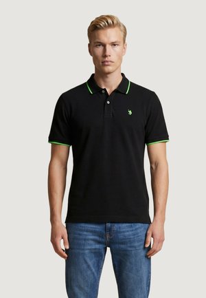 Hombre rubio con camiseta polo negra con ribete verde y pequeño logo, combinado con vaqueros azules, de pie contra un fondo claro.