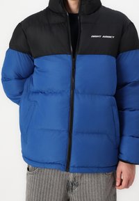 Veste matelass ée bleu et noir avec fermeture éclair, dotée d'un col haut, d'une texture rembourrée et du logo "NIGHT ADDICT" sur la poitrine.