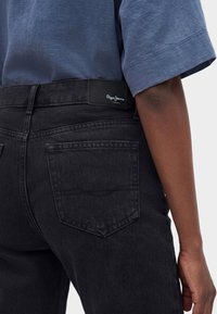 Άτομο που φοράει μαύρο τζιν από denim και μπλε κοντομάνικο πουκάμισο, δείχνοντας την πίσω τσέπη και μια μαύρη δερμάτινη ετικέτα με το λογότυπο "Pepe Jeans".