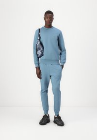 Felpa azzurra con polsini a costine, abbinata a pantaloni della tuta dello stesso colore. Borsa a tracolla nera con motivo, indossata a bandoliera. Sneakers nere.