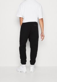 Schwarze, strukturierte Jogginghosen mit entspannter Passform, ausgestattet mit einem elastischen Bund und einer Gesäßtasche. Kombiniert mit weißen Sneakers und einem weißen Shirt.