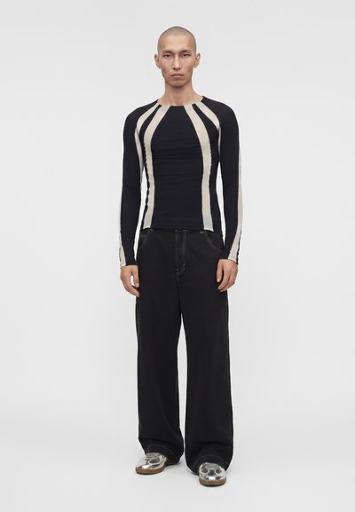 Άνδρας Ασιάτης στέκεται όρθιος φορώντας μια μακρυμάνικη μαύρη και μπεζ ριγέ μπλούζα, φαρδιά μαύρα παντελόνια και ασημένια αθλητικά παπούτσια σε λευκό φόντο.