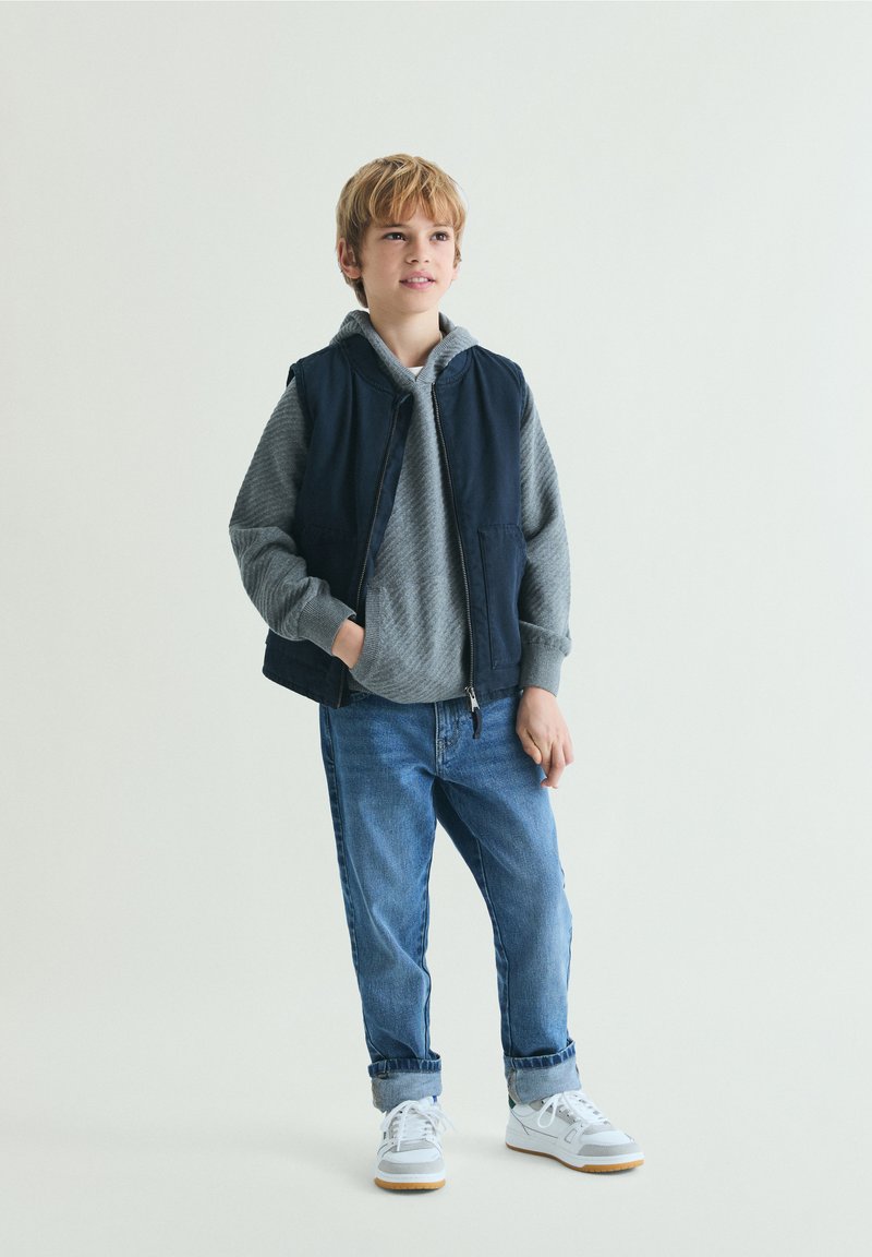 Garçon blond portant un sweat à capuche gris, un gilet bleu marine, un jean retroussé et des baskets blanches, debout avec une main dans la poche devant un fond uni.