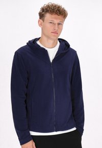 Felpa con zip color navy realizzata in tessuto morbido, dotata di cappuccio, maniche lunghe e orlo dritto. Indossata sopra una maglietta bianca.