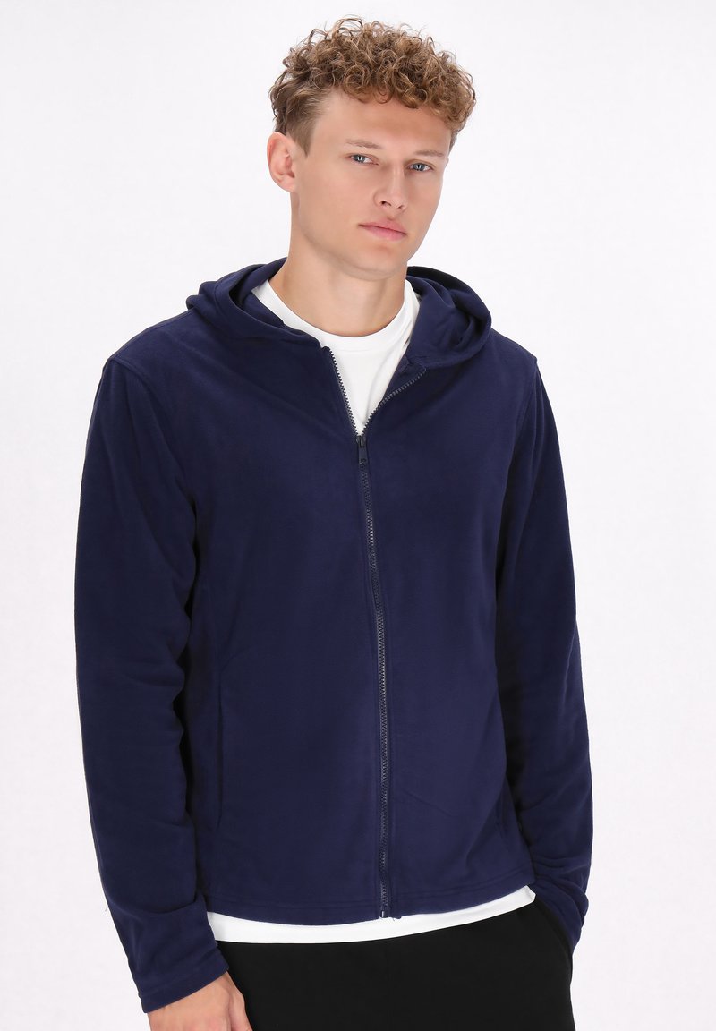 Felpa con zip color navy realizzata in tessuto morbido, dotata di cappuccio, maniche lunghe e orlo dritto. Indossata sopra una maglietta bianca.
