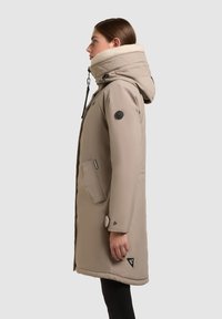 Cappotto beige imbottito con cappuccio foderato in ecopelliccia, texture a diamond diagonale, tasche laterali e patch con logo sulla manica.