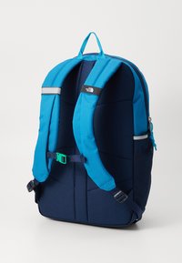 Mochila com um exterior azul e detalhes em azul marinho, alças acolchoadas, acabamentos refletivos e um bolso lateral em malha. Tecido durável e forma estruturada.