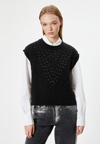 Schwarzes, kurzärmliges Strickwesten mit einem Zopfmuster und Perlenakzenten in V-Form. Über einem weißen Hemd mit Kragen getragen.