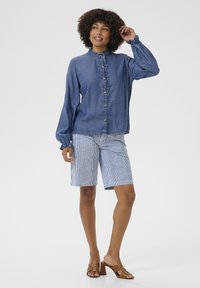 Denim overhemd met knopen, voorzien van een gerimpelde kraag en manchetten, gecombineerd met gestreepte shorts. Model draagt bruin blokhak sandalen. Helder witte achtergrond.