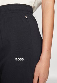 Melni sporta bikses ar elastīgu vidukli, balta „BOSS” logotipa apdare, mīksta tekstūra un neliels kontrastējošās krāsas zīmola elements.