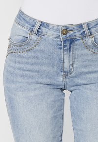 Jeans de mezclilla azul claro con acentos de tachuelas doradas a lo largo de los bolsillos, un diseño ajustado, cinco bolsillos y un cierre de botón metálico en la cintura.