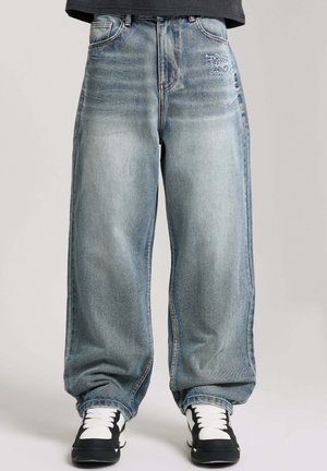 Lässig geschnittene, ausgewaschene blaue Jeans mit gesticktem Logo auf der rechten Tasche, getragen mit schwarzen und weißen Turnschuhen vor neutralem Hintergrund.