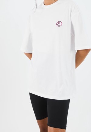 Persoon die een wit oversized t-shirt draagt met een kleine roze en zwarte smiley patch en zwarte knielange shorts.