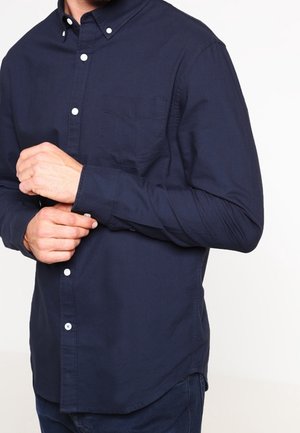 Mann, der ein dunkelblaues Hemd mit langen Ärmeln trägt, richtet die Manschette seines linken Handgelenks vor einem einfarbigen Hintergrund.