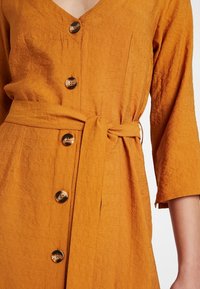 Senapsgul linneklänning med djup V-ringning, fem dekorativa knappar och bältet knutet i midjan. Texturerat tyg med 3/4-långa ärmar.