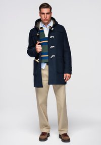 Marineblå uld duffelcoat med hætte, med trætoggle lukninger, to forlommer, lagdelt over en grønstribet sweater og beige bukser.