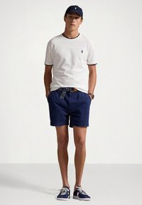 Λευκό κοντομάνικο t-shirt με μαύρο περιθώριο, σκούρα μπλε σορτς και σκούρα μπλε αθλητικά παπούτσια. Αξεσουάρ περιλαμβάνουν σκούφο σε σκούρο μπλε και ριγωτή ζώνη.