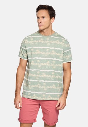 Hombre con camiseta verde y blanca con patrón de palmeras y pantalones cortos color coral enrollados, de pie con las manos relajadas a los lados.