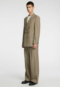 Bež dvoprsni blazer s črnimi gumbi, kombiniran s širokimi hlačami. Prilagojena oblika, gladka tekstura in subtilni detajli šivov.