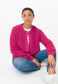 Cardigan de punto fucsia con cuello redondo, cierre de botones y patrón texturizado. Combinado con jeans azul claro y calzado blanco.