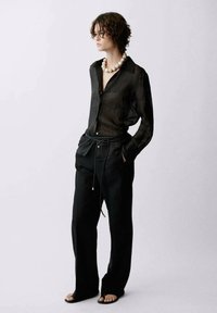 Black sheer button-up shirt with a collar, paired with loose black trousers and flat sandals. Necklace features large white beads.