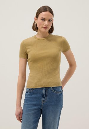 Joven con cabello corto que lleva pendientes de aro dorados, una blusa ajustada de manga corta color mostaza y jeans azules de cintura alta, posando frente a un fondo liso.