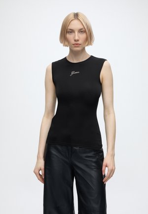 Jeune femme aux cheveux blonds courts portant un haut noir sans manches ajusté et un pantalon en cuir noir ample, debout devant un fond uni.