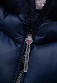 Giacca puffer blu navy con zip frontale, dotata di un tirante argento con l'etichetta "ombre performance". Tessuto liscio e lucido con texture trapuntata.