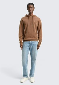 Felpa marrone con tasca frontale, cappuccio con coulisse e un piccolo logo. Abbinata a jeans azzurri chiari e sneakers bianche. Design semplice.