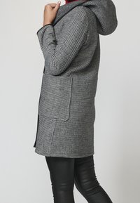 Cappotto a motivo pied-de-poule in bianco e nero, dotato di cappuccio, maniche lunghe, due tasche laterali e orlo dritto.