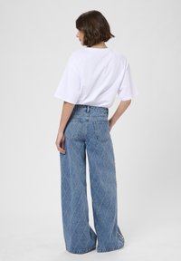 Žena s krátkymi hnedými vlasmi má na sebe voľné biele tričko a džínsy s vysokým pásom a širokými nohavicami s diamantovým šitím a perlovými detailmi po stranách.