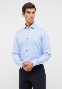Chemise de couleur bleu clair avec un col écarté, des manches longues et des détails de bouton marron, présentant un motif texturé subtil.