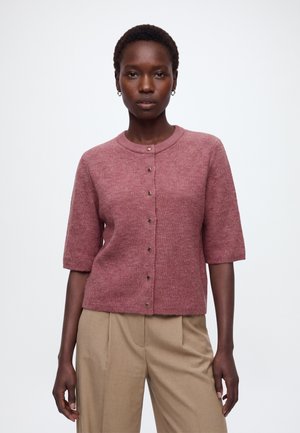 Kvinde iført en støvet lyserød kortærmet cardigan med knapper og beige bukser med høj talje, stående mod en almindelig hvid baggrund.