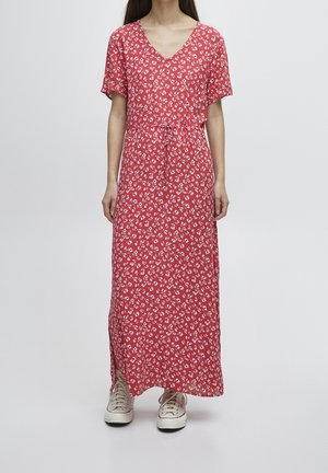 Femme portant une robe maxi rouge à fleurs à manches courtes avec une taille à cordon de serrage et des baskets blanches, debout devant un fond uni.