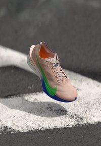 Világos rózsaszín futócipő zöld és kék részletekkel, ami aszfalt felett lebeg fehér útjelölésekkel.