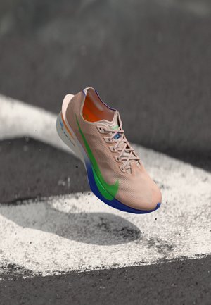 Gaiši rozā skriešanas apavi ar zaļiem un ziliem akcentiem, kas peld virs asfalta ar baltu ceļa marķējumu.