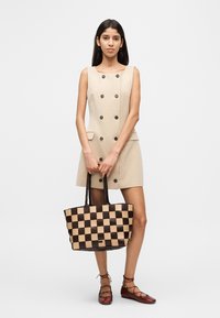 Beiges Kleid mit doppelreihigen Knöpfen, kombiniert mit einer braun-beigen karierten Tragetasche und braunen flachen Schuhen. Insgesamt glatte Textur.