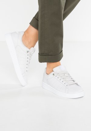 Persona con indosso scarpe da ginnastica bianche allacciate e pantaloni verde oliva arrotolati, in piedi su un pavimento bianco davanti a uno sfondo bianco.