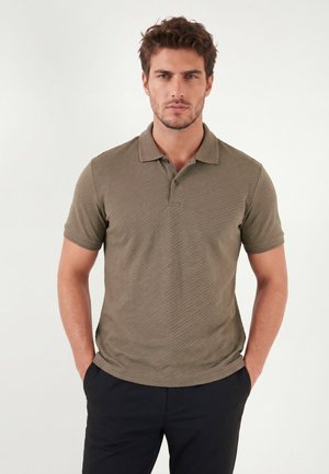 Jonge man met kort bruin haar, gekleed in een bruin poloshirt met korte mouwen en een zwarte broek, staand met handen in de zakken tegen een effen achtergrond.