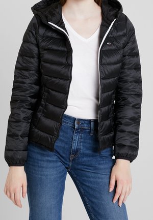 Kvinde iført en sort dunjakke med hætte, hvid V-hals t-shirt og blå jeans mod en neutral baggrund.