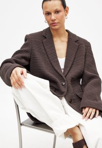 Brauner strukturierter Blazer mit Revers, zwei Knöpfen, getragen über einem weißen Hemd und kombiniert mit weißen Hosen. Sitzt auf einem Stuhl.