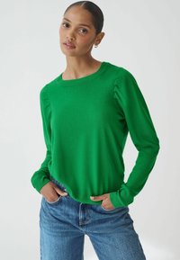 Grøn striktrøje med lange ærmer, ribbede manchetter og diskrete rynkedetaljer på skuldrene, parret med blå denimjeans.