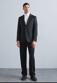 Černý tailor-made oblek s klopovým límcem a jedním knoflíkem, v kombinaci s bílou košilí a lesklými černými botami, stojící na jednobarevném pozadí.