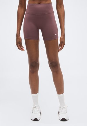 Atletisch persoon staat, draagt mauve hoog geplaatste korte broek, witte sportsokken en witte vetersneakers tegen een effen lichte achtergrond.