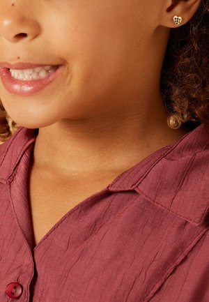 Bordeaux shirt met een gestructureerde afwerking en kraag, met een knoopsluiting. Het model heeft een kleine gouden hartvormige oorbel met kleurrijke accenten.