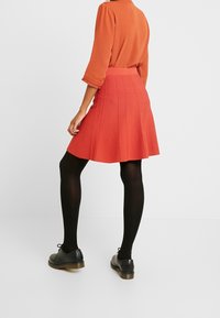 Jupe évasée rouge avec une taille ajustée, présentant des coutures verticales et une finition texturée. Portée avec des collants et des chaussures noirs.