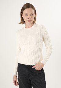 Krémový svetr s pleteným vzorem, kulatým výstřihem, přiléhavými rukávy a žebrovaným lemem. Nosit s tmavými džíny, které ukazují texturovanou látku.