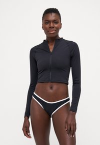 Black cropped rash guard with long sleeves and a front zipper, paired with black bikini bottoms featuring a white trim. Smooth fabric texture.